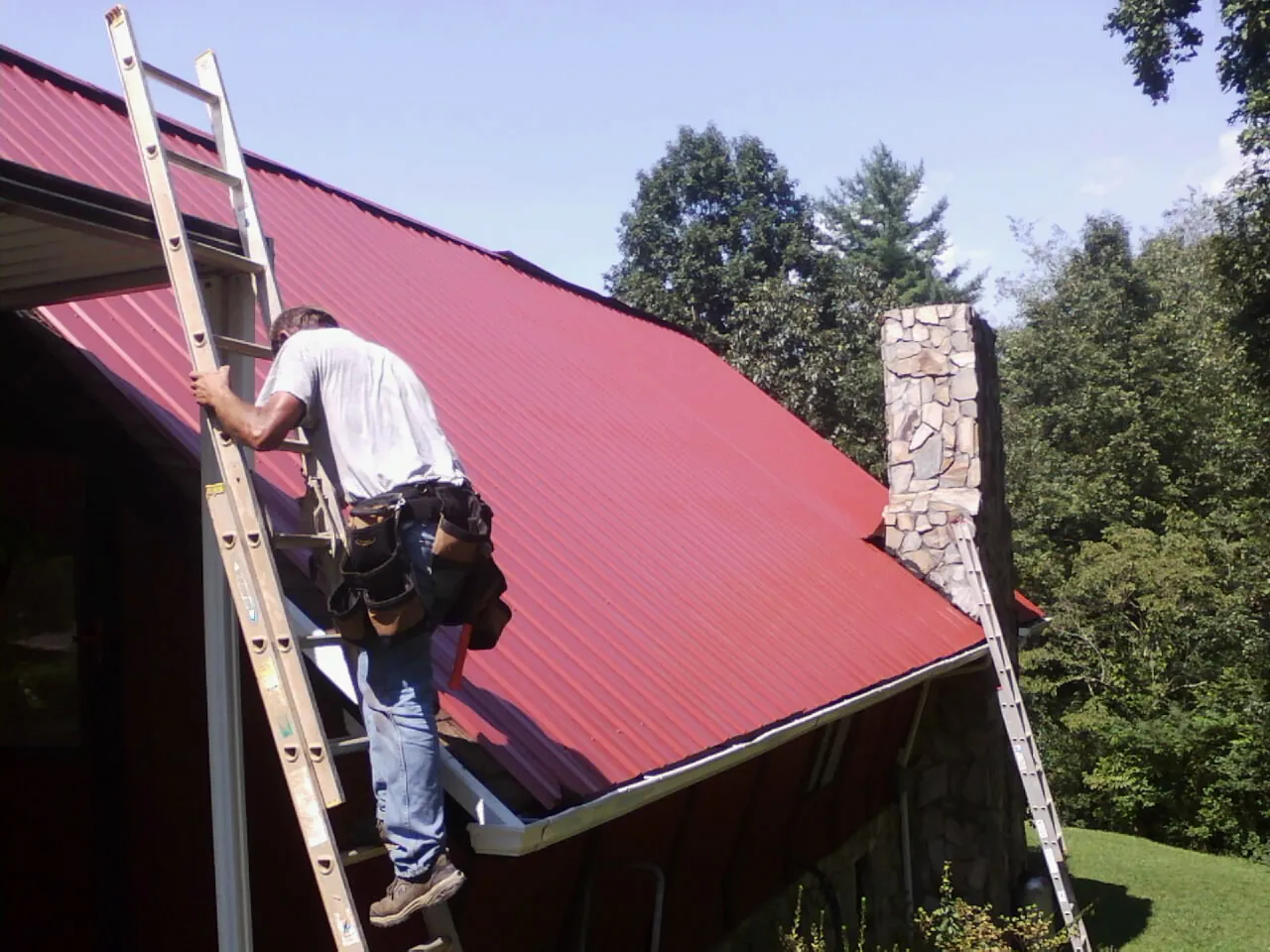 Roofer on a ladder beside a red metal roof and stone chimney — Soffit & Fascia Repair by Roofing Brothers in Elsinore